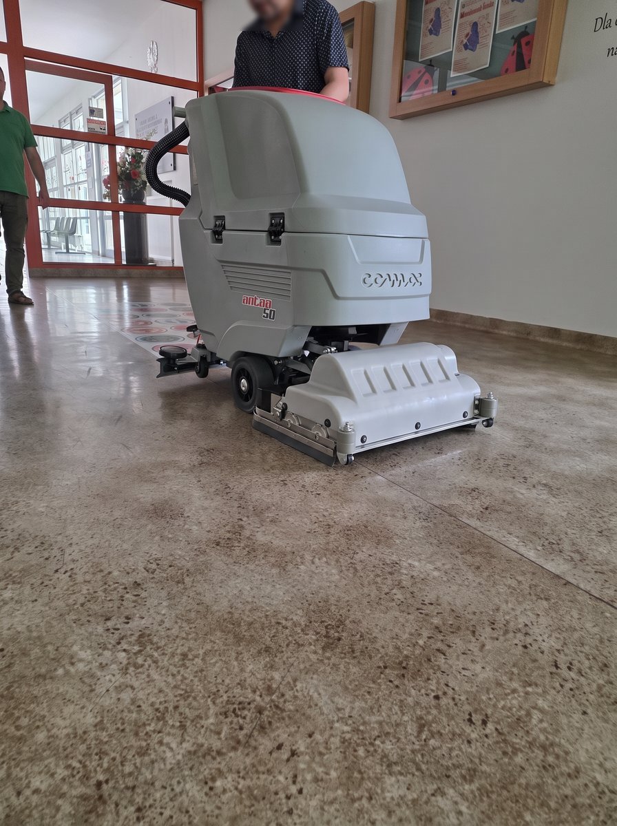 Antea 50 BTS prowadzona przez operatora na korytarzu szkoły — głowica z walcami, skala maszyny na tle człowieka