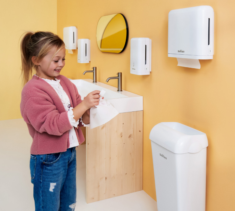 Dziewczynka w toalecie wyciera ręce ręcznikiem papierowym po umyciu dłoni w tle widać gamę produktów satino by wepa dozowniki do mydła w płynie i piance