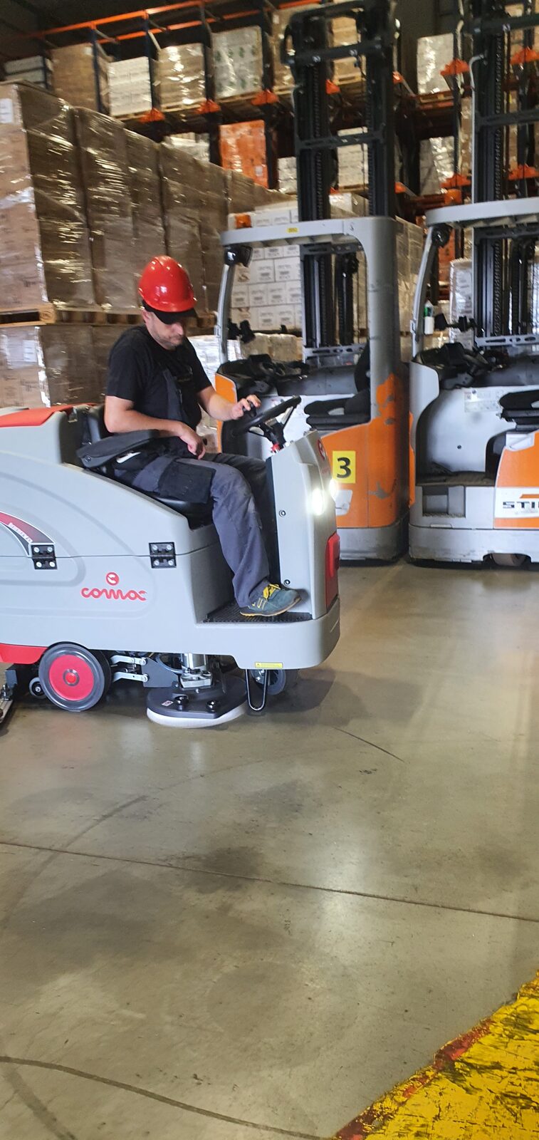 Comac Innova 85 B sprząta halę magazynową obok wózka widłowego